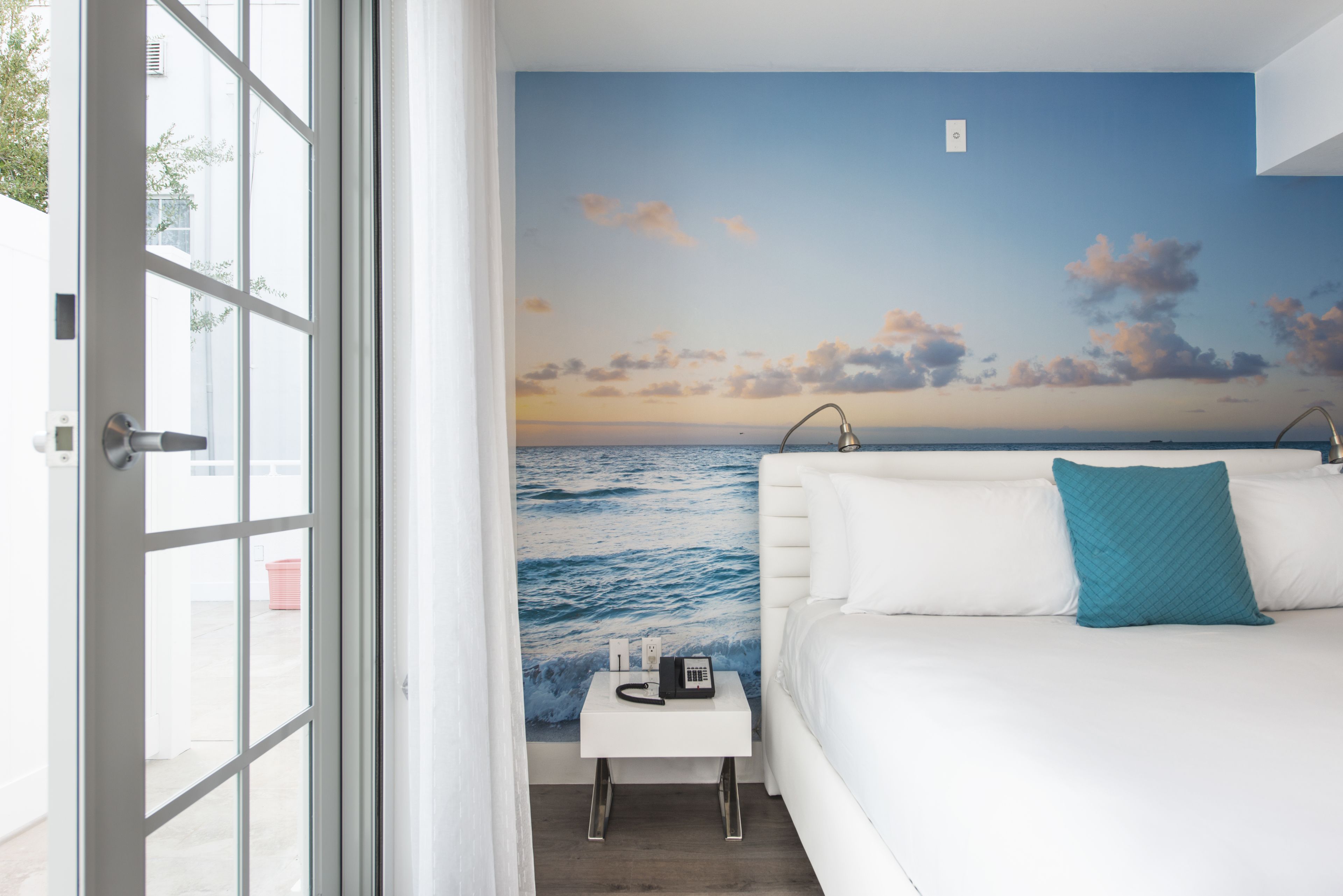 Un dormitorio con vista al océano a través de una ventana, una cama blanca y una mesita de noche pequeña con un teléfono y una taza.