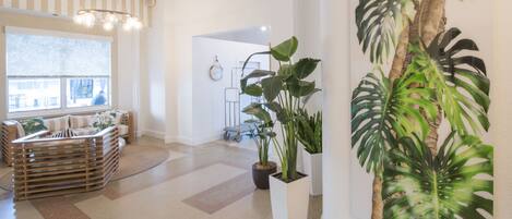 Een ruime kamer met een groot raam, een moderne kroonluchter, een zithoek met een bank en stoelen, en diverse potplanten.