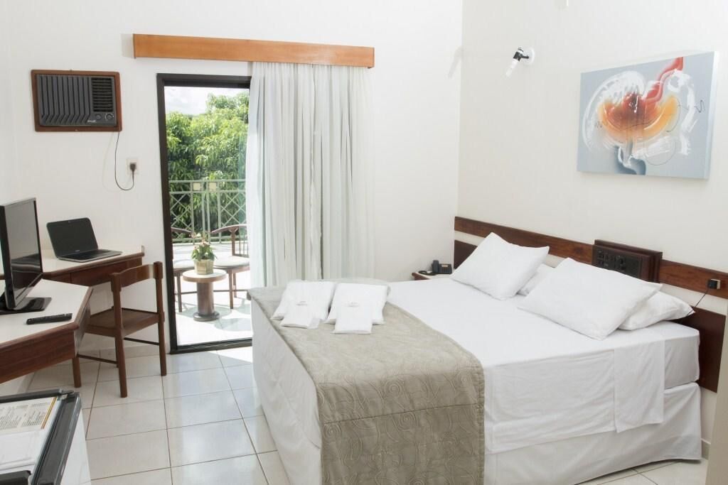 A hotel room with a bed, desk, laptop, and a view of greenery outside.