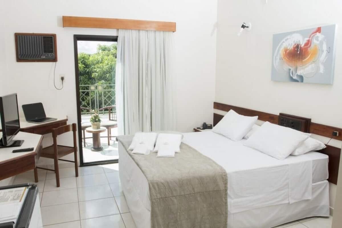 A hotel room with a bed, a desk with a laptop, a chair, a small table with a plant, and a balcony with a view of greenery.