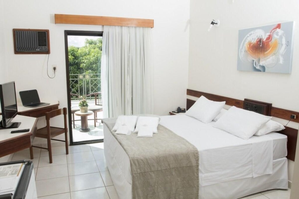 A hotel room with a bed, a desk with a laptop, a chair, a small table with a plant, and a balcony with a view of greenery.