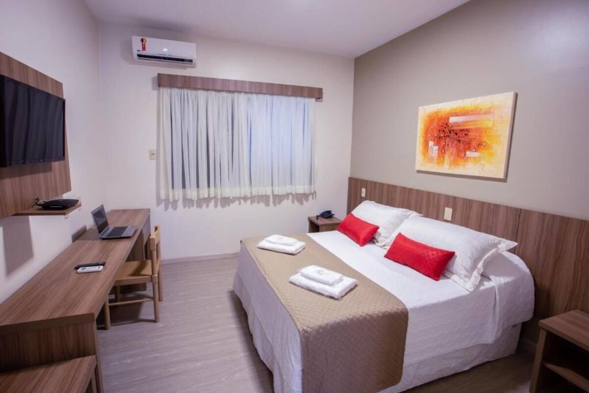 A hotel room with a bed, a desk, a chair, an air conditioning unit, a window with curtains, and a painting on the wall.