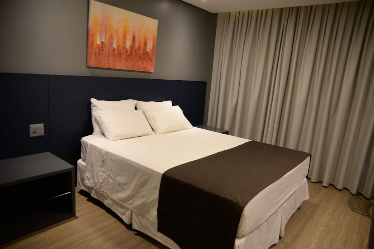 A hotel room with a large bed, a nightstand, a wall-mounted painting, and a window with curtains.