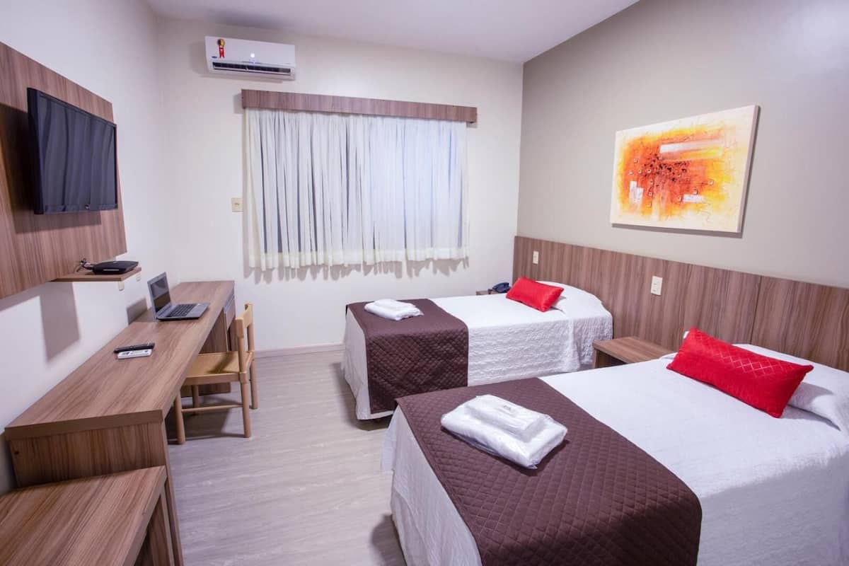 A hotel room with two beds, a desk, a chair, a TV, and an air conditioning unit.