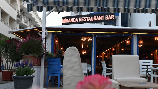 An outdoor seating area with a restaurant bar named 'Miranda Restaurant Bar'. There are potted plants, outdoor chairs, and tables.