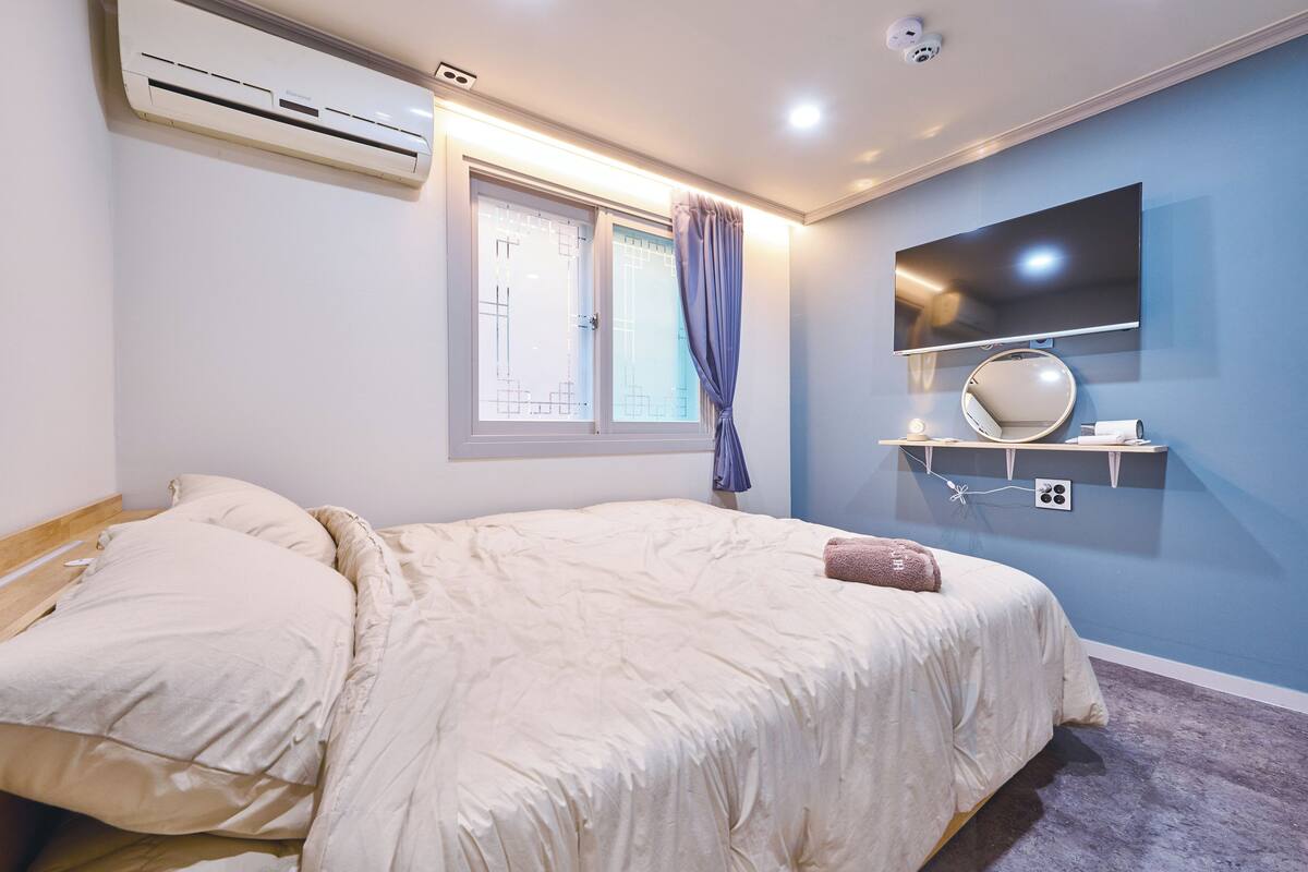 A bedroom with a bed, air conditioning unit, window with curtains, a mounted TV, and a small shelf with a mirror and a cup.