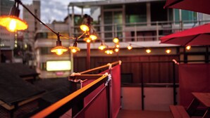 Une terrasse sur le toit, agrémentée de guirlandes lumineuses et de parasols rouges, d’où l’on peut admirer un immeuble aux grandes fenêtres.