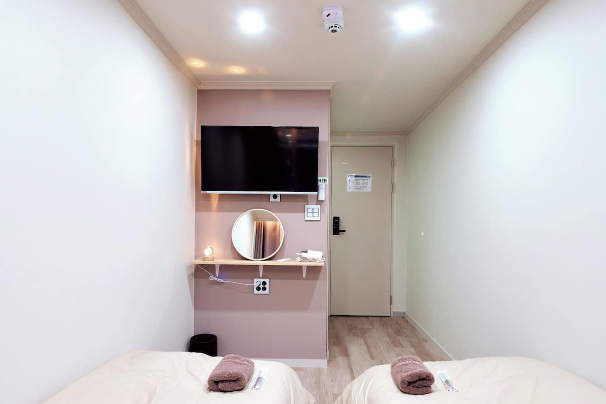 A hotel room with two beds, a TV mounted on the wall, a mirror, a small shelf with a lamp, and a door leading to another room.