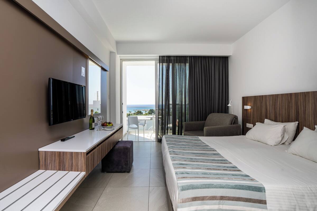 A modern hotel room with a large bed, a desk with a flat-screen TV, a seating area with a chair, and a balcony with a view.