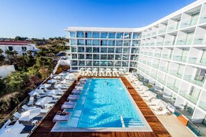 Un hotel moderno con una amplia piscina cubierta rodeada de tumbonas y sombrillas, ventanas amplias y vistas del paisaje circundante.