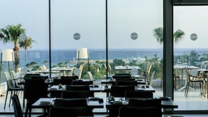 Ein leeres Restaurant mit Tischen und Stühlen, große Fenster mit Blick auf das Meer und Gebäude.