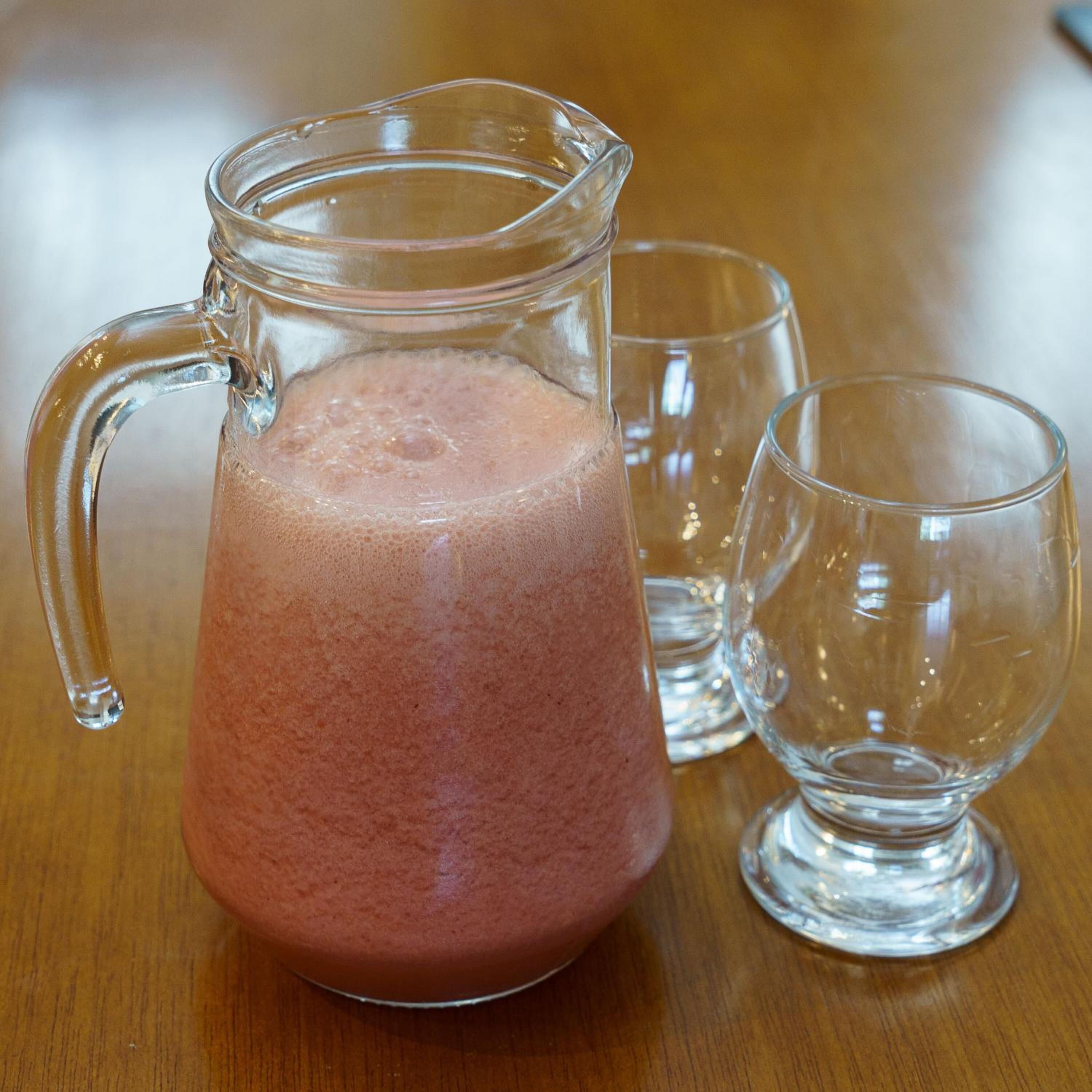 A glass pitcher filled with a pink beverage, accompanied by two empty glasses on a wooden table.