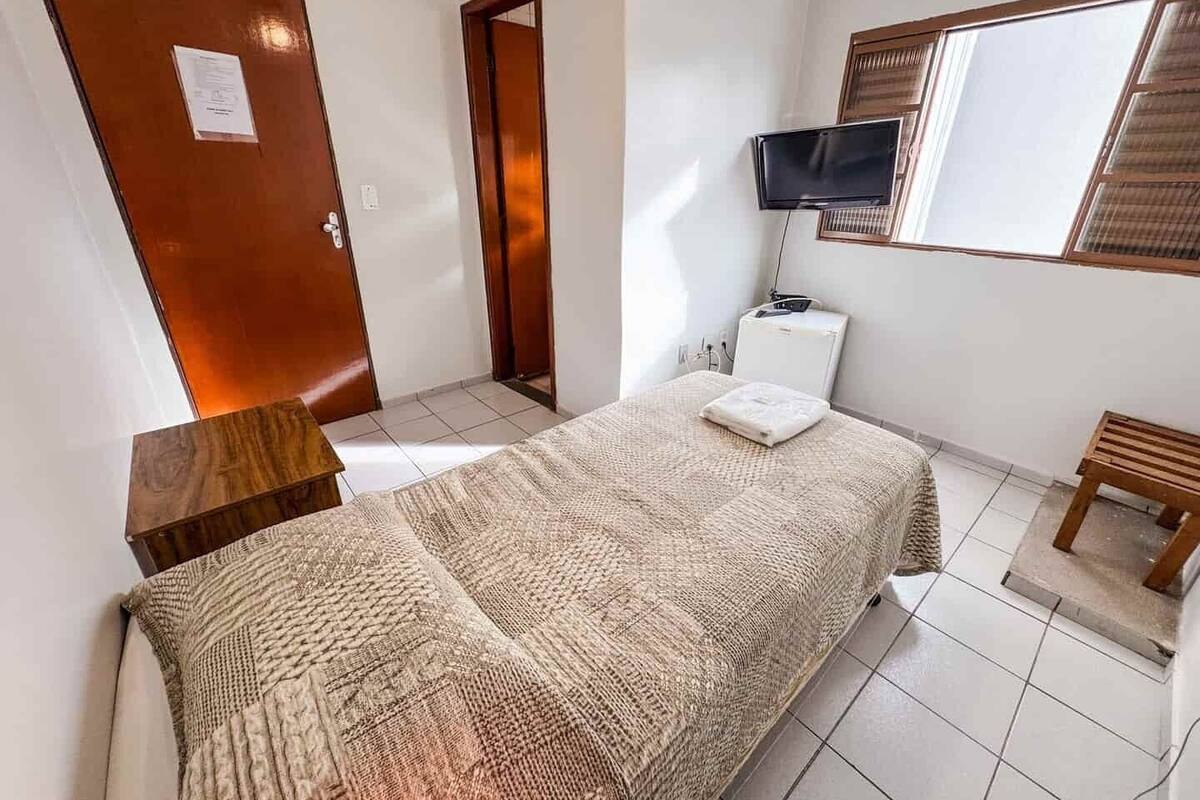 A bedroom with a bed, a wooden nightstand, a TV, and a window.