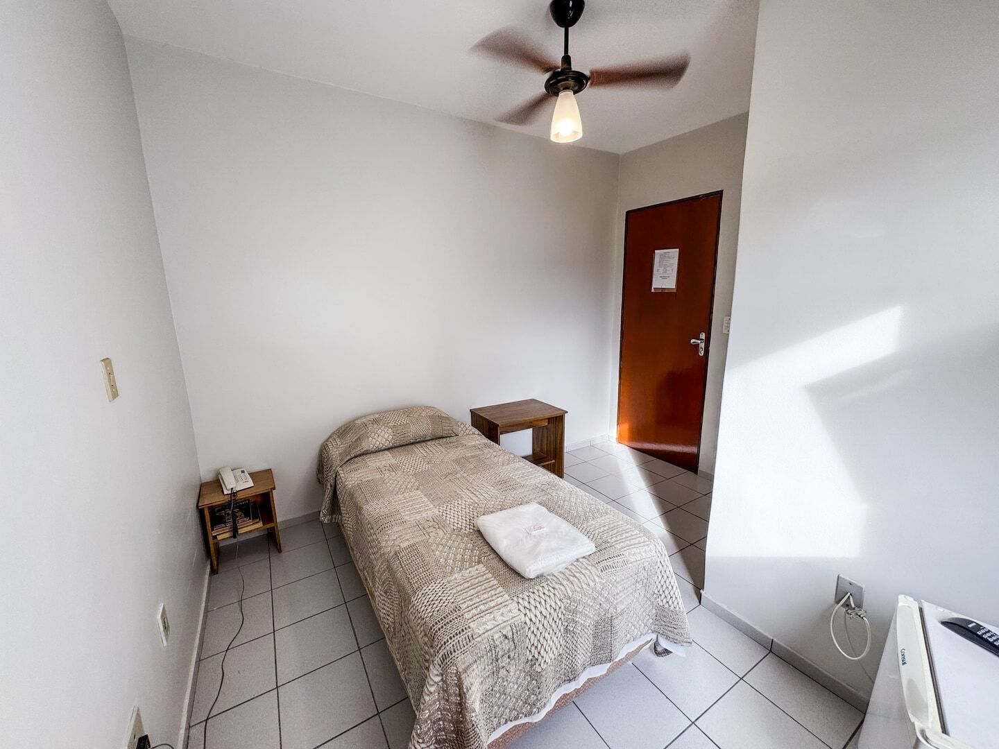 A small, clean room with a bed, a nightstand, a wooden door, and a ceiling fan.