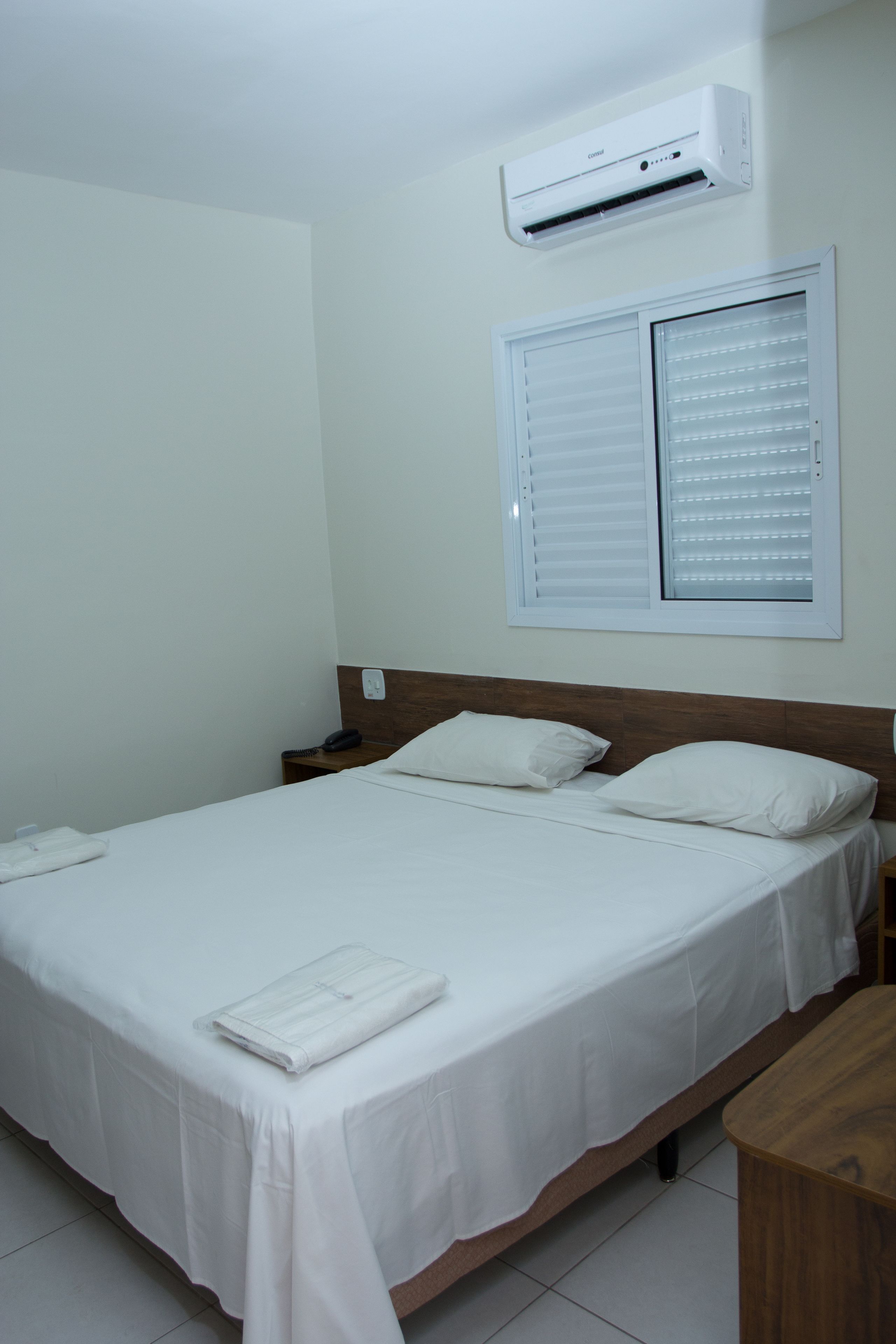 Una cama bien hecha con sábanas blancas, un cabecero de madera, una mesita de noche con lámpara y un aire acondicionado de pared.
