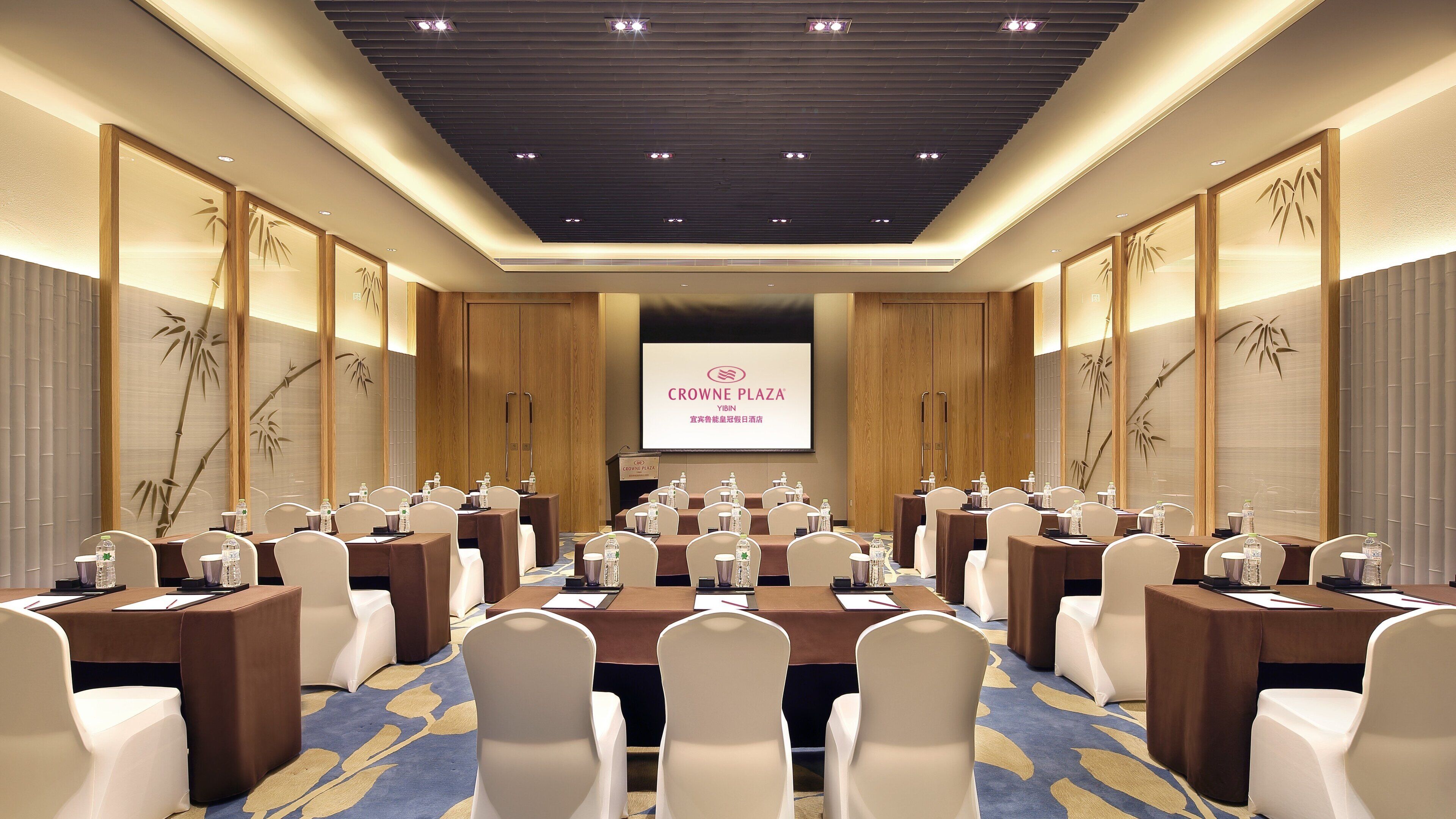A conference room with multiple tables and chairs, a large screen displaying the Crowne Plaza Yibin logo, and wooden paneling with bamboo designs.