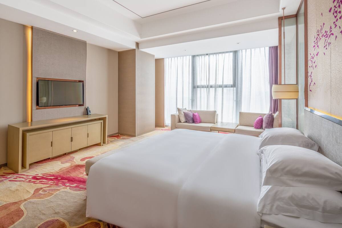 A hotel room with a large bed, a TV mounted on the wall, a sofa with cushions, and a window with curtains.