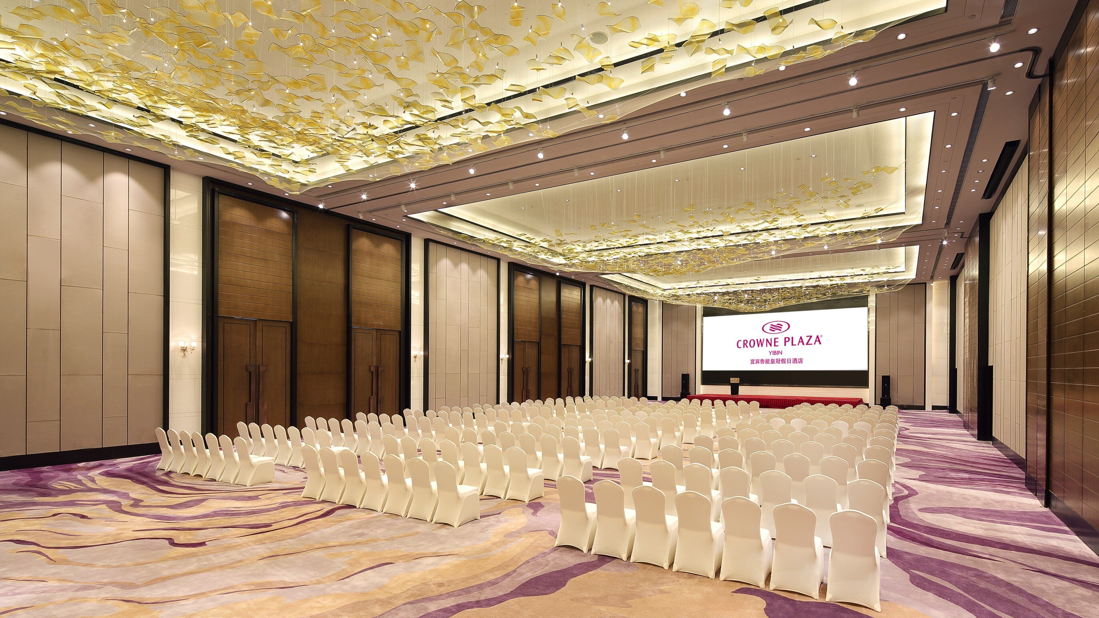 A conference room with rows of white chairs, a large screen displaying the Crowne Plaza Yibin logo, and a patterned carpet.