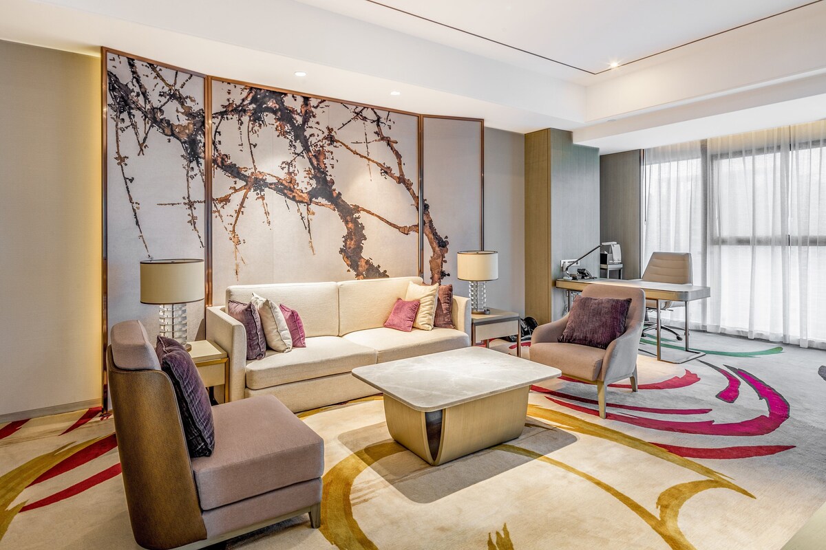 A modern hotel room with a sofa, armchair, coffee table, and a large abstract painting on the wall.