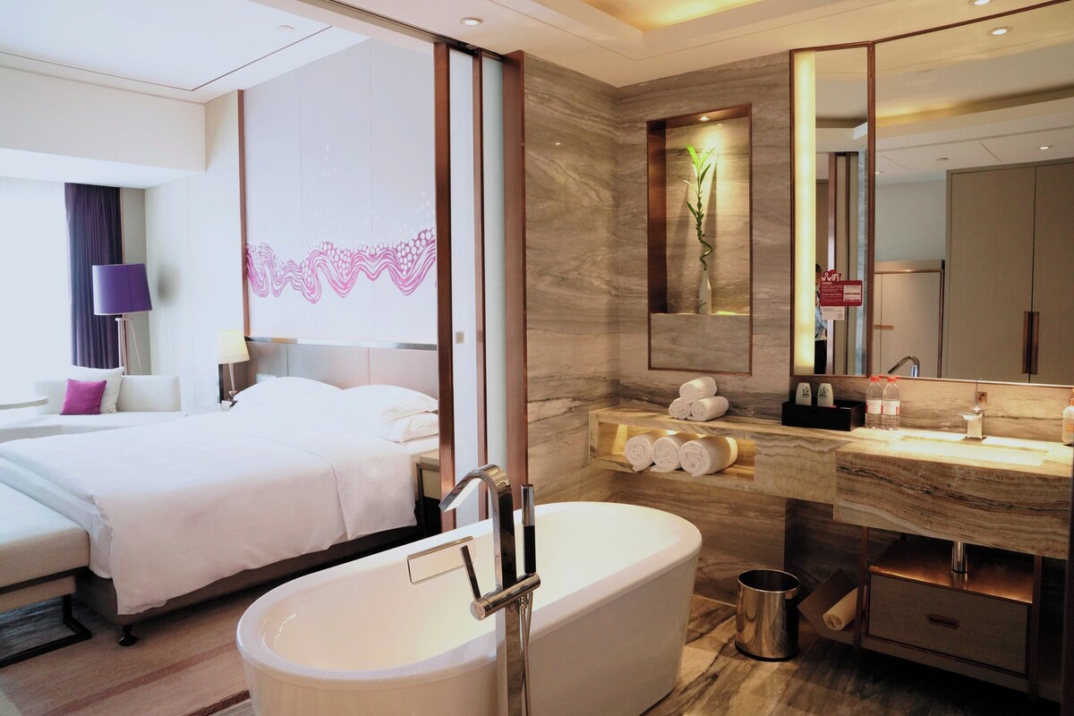 A modern hotel room with a large bed, a separate bathroom with a bathtub, a vanity with a mirror, and a wall-mounted toilet.
