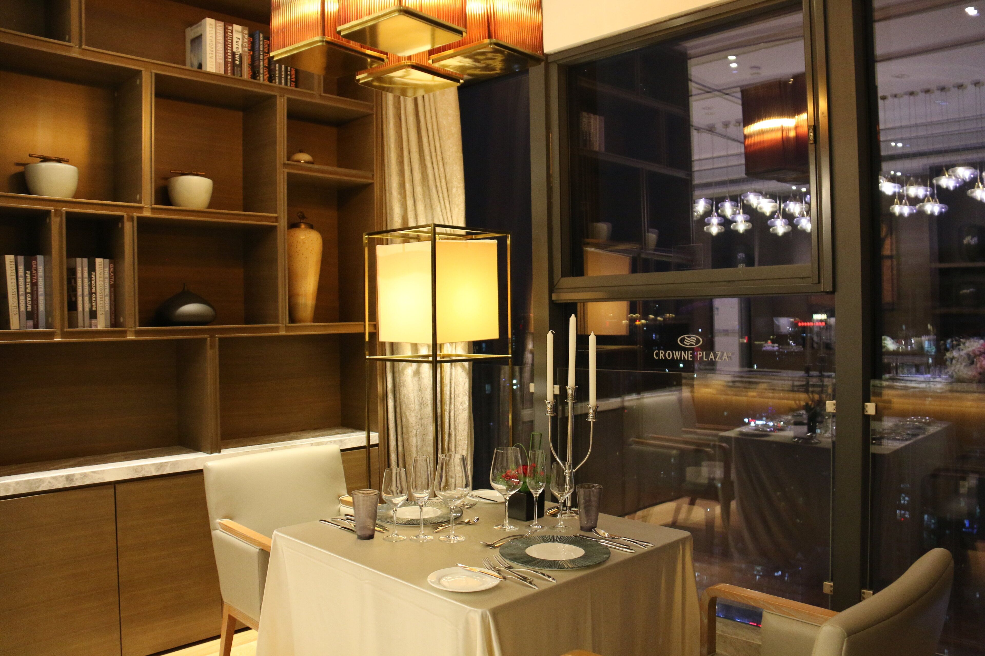 A dining area with a table set for four, a bookshelf with books and decorative items, and a view of the city through a large window.