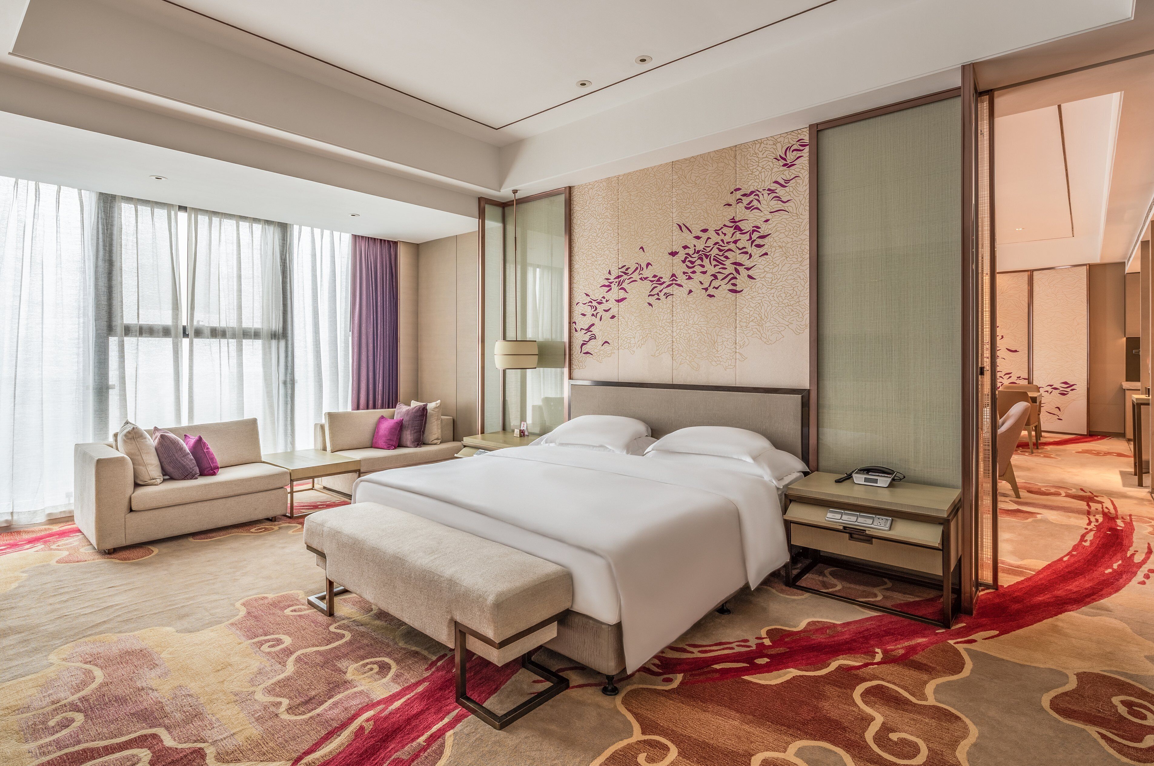 Quarto de hotel moderno com cama grande, sofá, escrivaninha e uma janela ampla com cortinas leves.