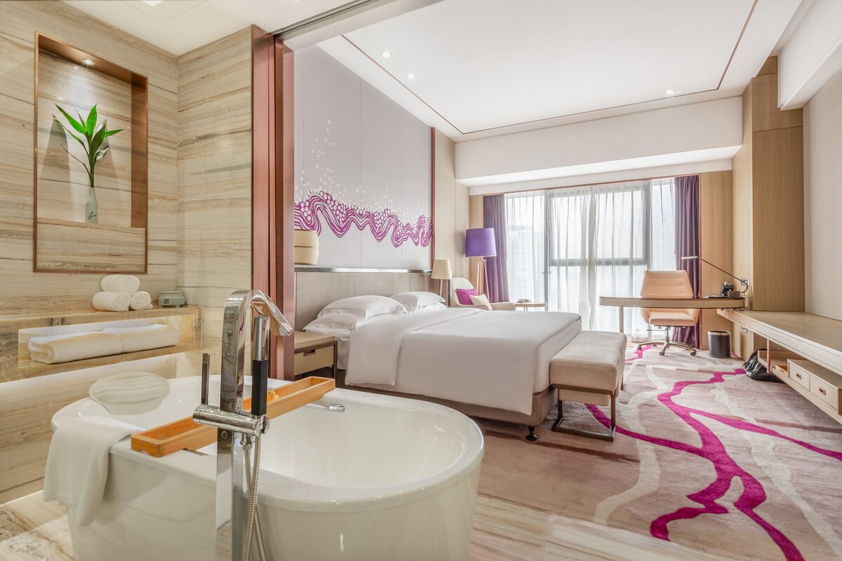 A modern hotel room with a large bed, a sitting area, a desk, and a bathroom with a bathtub and a sink.