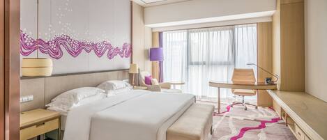 A modern hotel room with a large bed, a desk, a chair, and a view of the city through large windows.