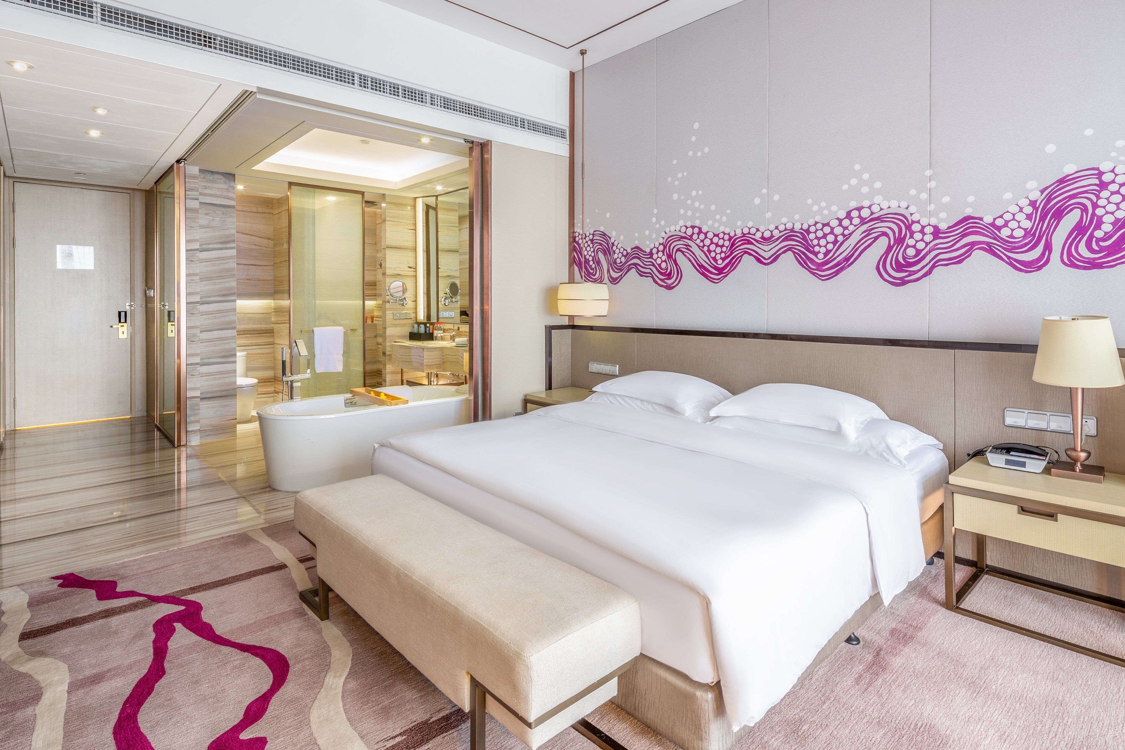 Habitación de hotel moderna con una cama grande, un banco, un escritorio y un baño visible a través de una puerta abierta.