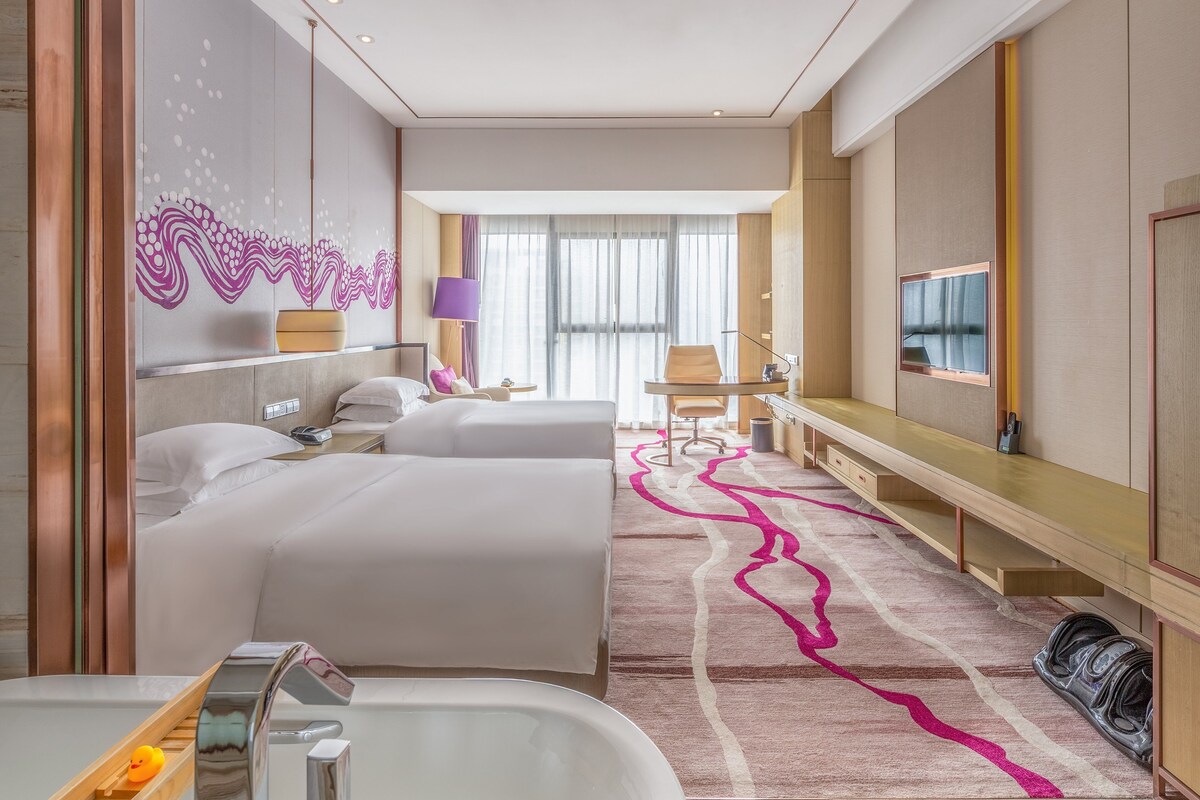A hotel room with two beds, a desk, a TV, and a bathroom with a bathtub and shower.