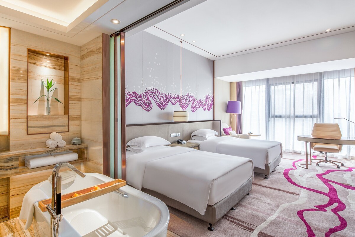 A hotel room with two beds, a bathtub, a desk, and a chair.