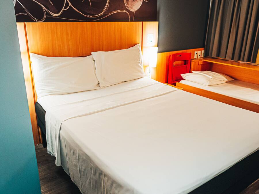 A hotel room with a large bed, a smaller bed, a red chair, and a nightstand with a lamp.