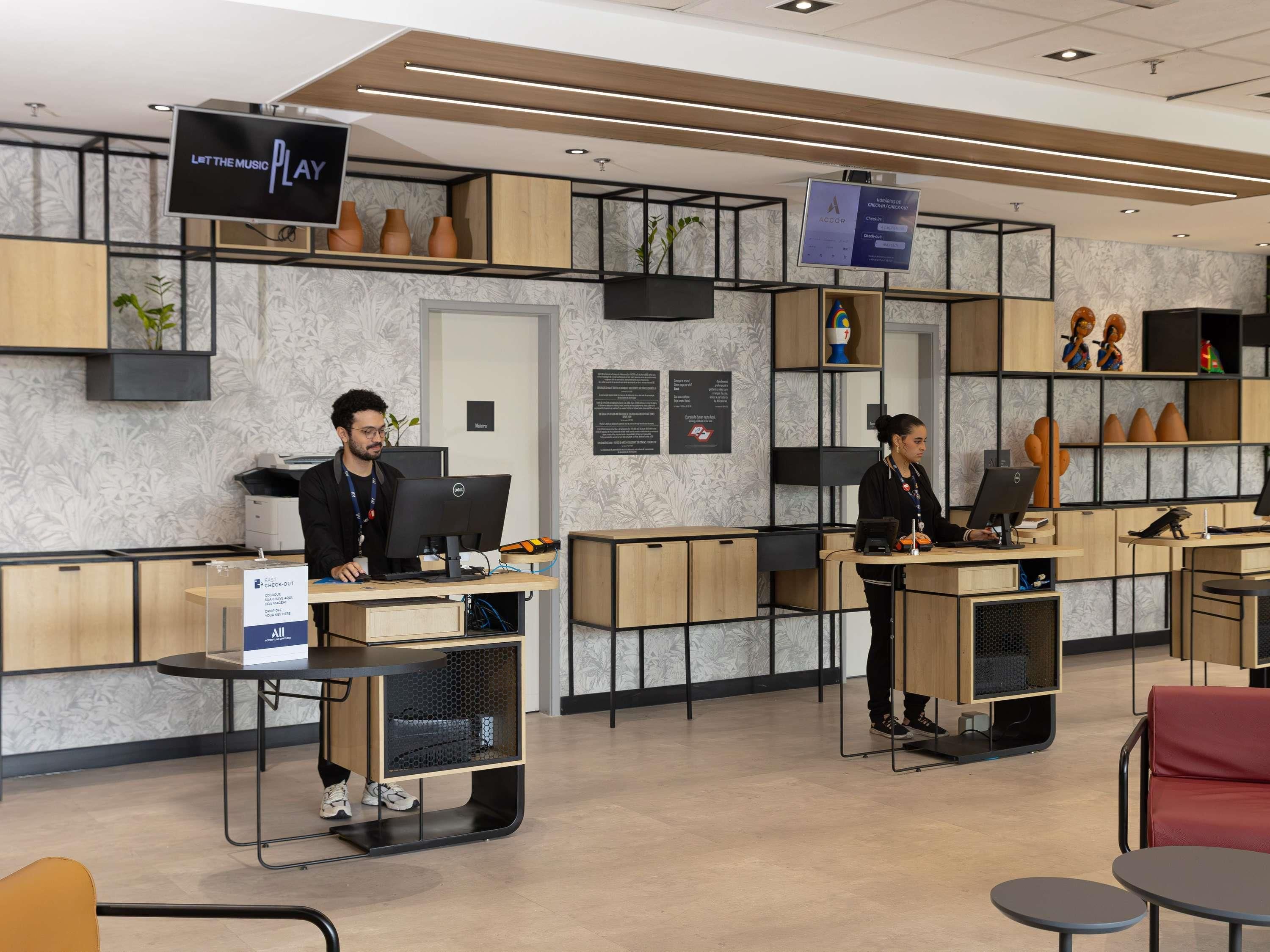 A modern reception area with two receptionists at desks, a digital screen displaying 'LET THE MUSIC PLAY', and a sign for 'ACCOR CHECK-IN'.