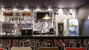 Un área de bar moderna con un mural abstracto en blanco y negro, una máquina de café y asientos con sillas rojas y blancas.