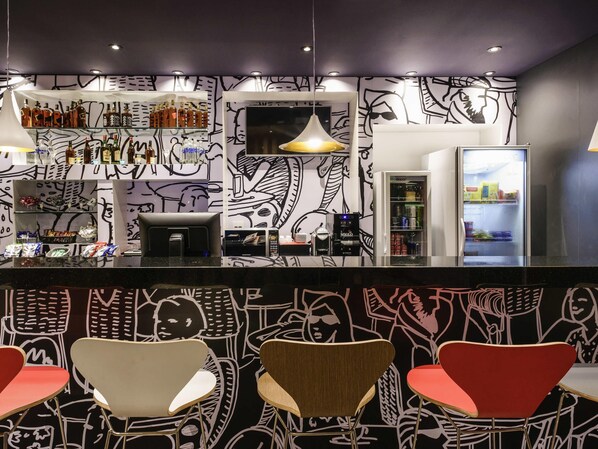 Un área de bar moderna con un mural abstracto en blanco y negro, una máquina de café y asientos con sillas rojas y blancas.