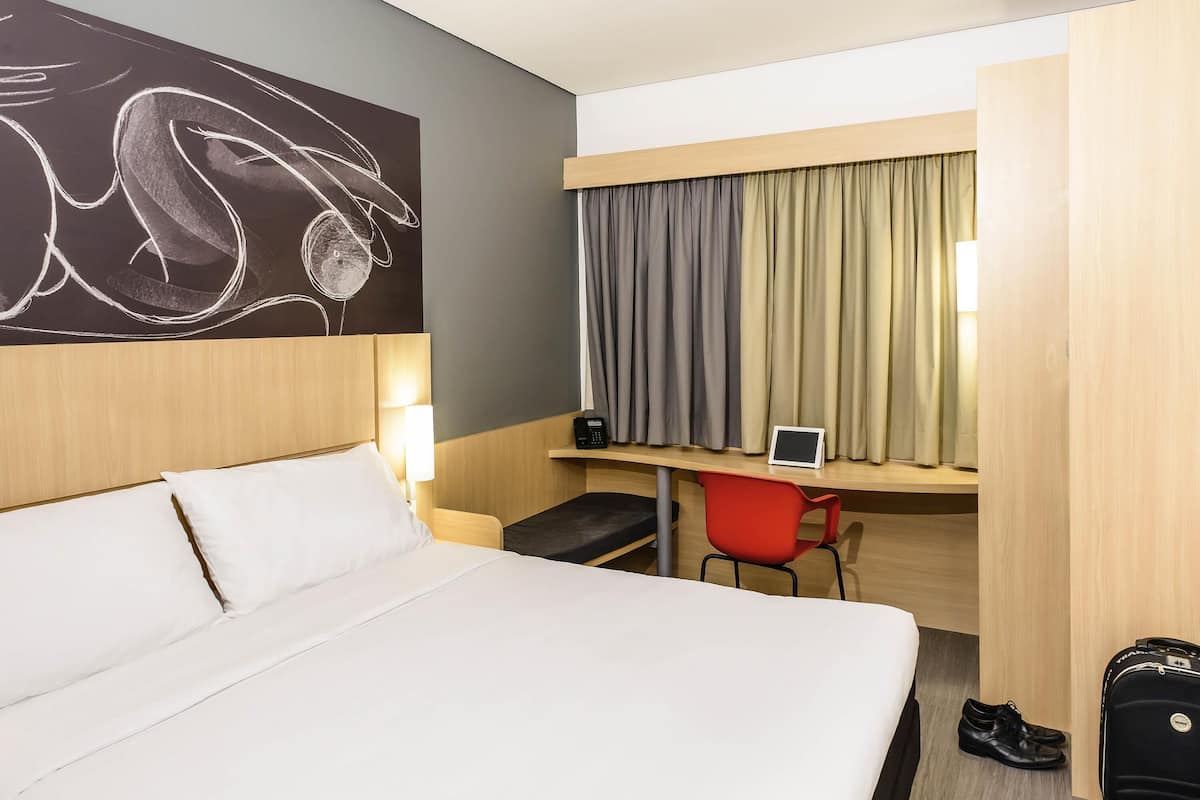 A hotel room with a bed, a desk with a chair, a phone, a tablet, and a suitcase.