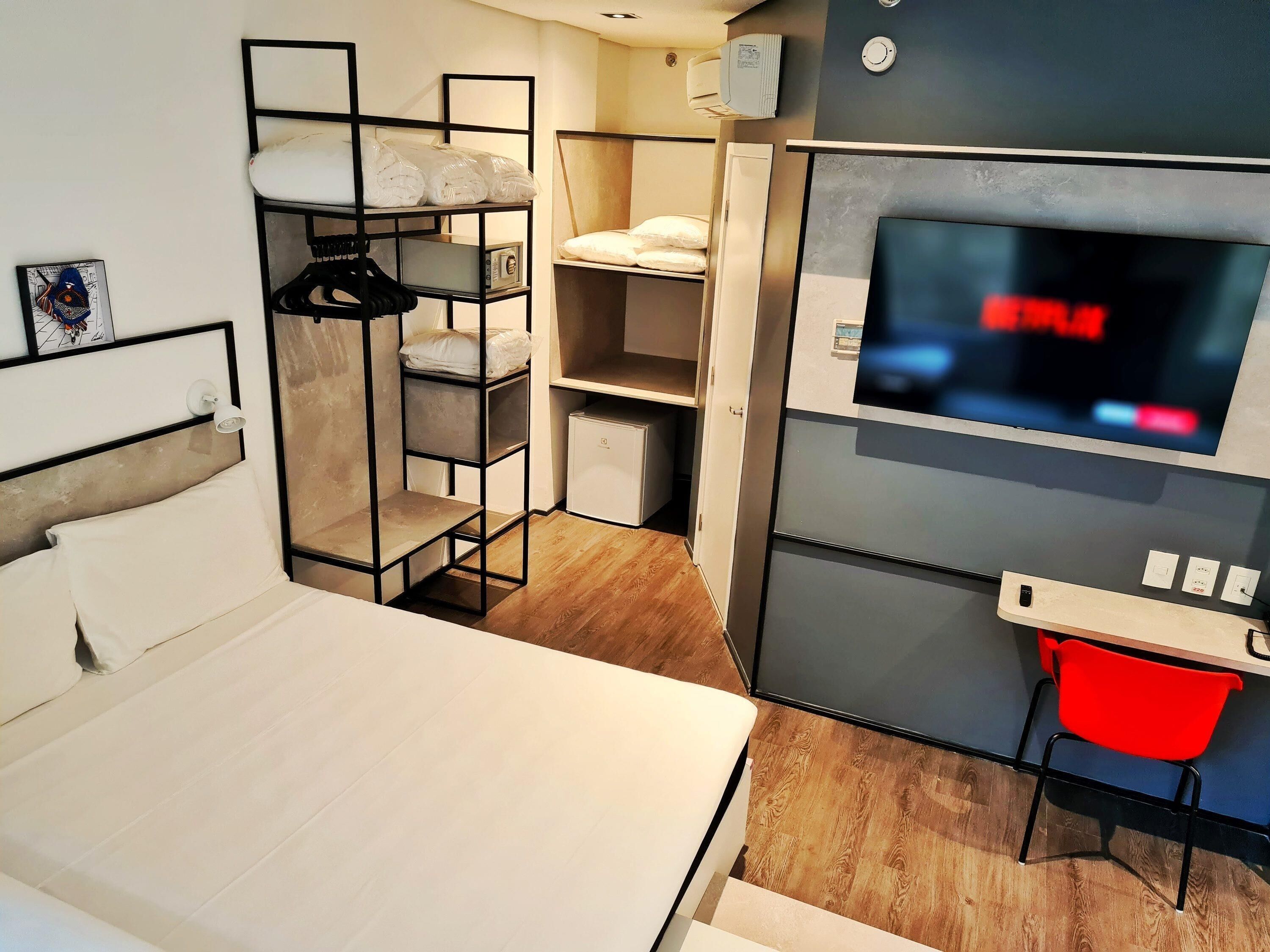 Habitación de hotel compacta con cama, televisor, escritorio pequeño y armario con estantes.