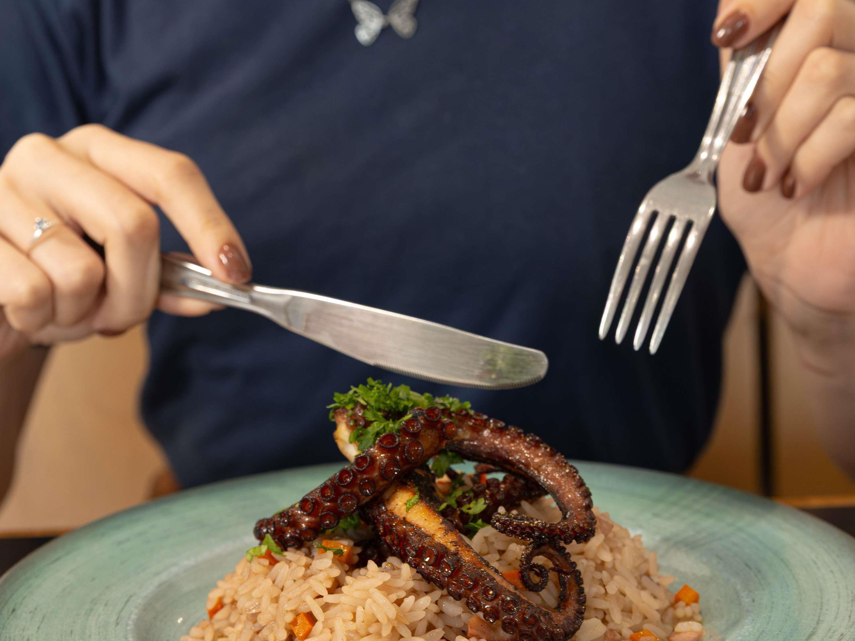 A person holding a fork and knife, about to eat a meal with octopus tentacles on top of rice.