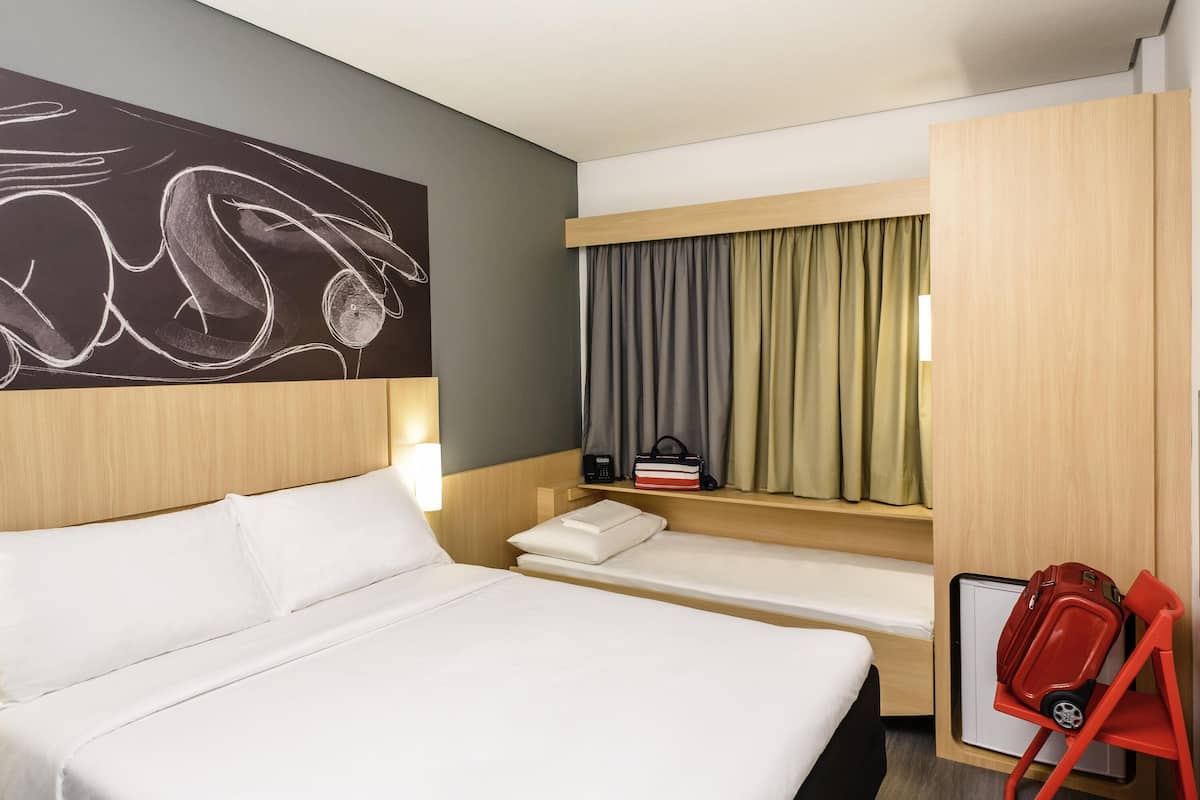 A hotel room with a large bed, a smaller bed, a desk with a chair, a window with curtains, and a wall art piece.
