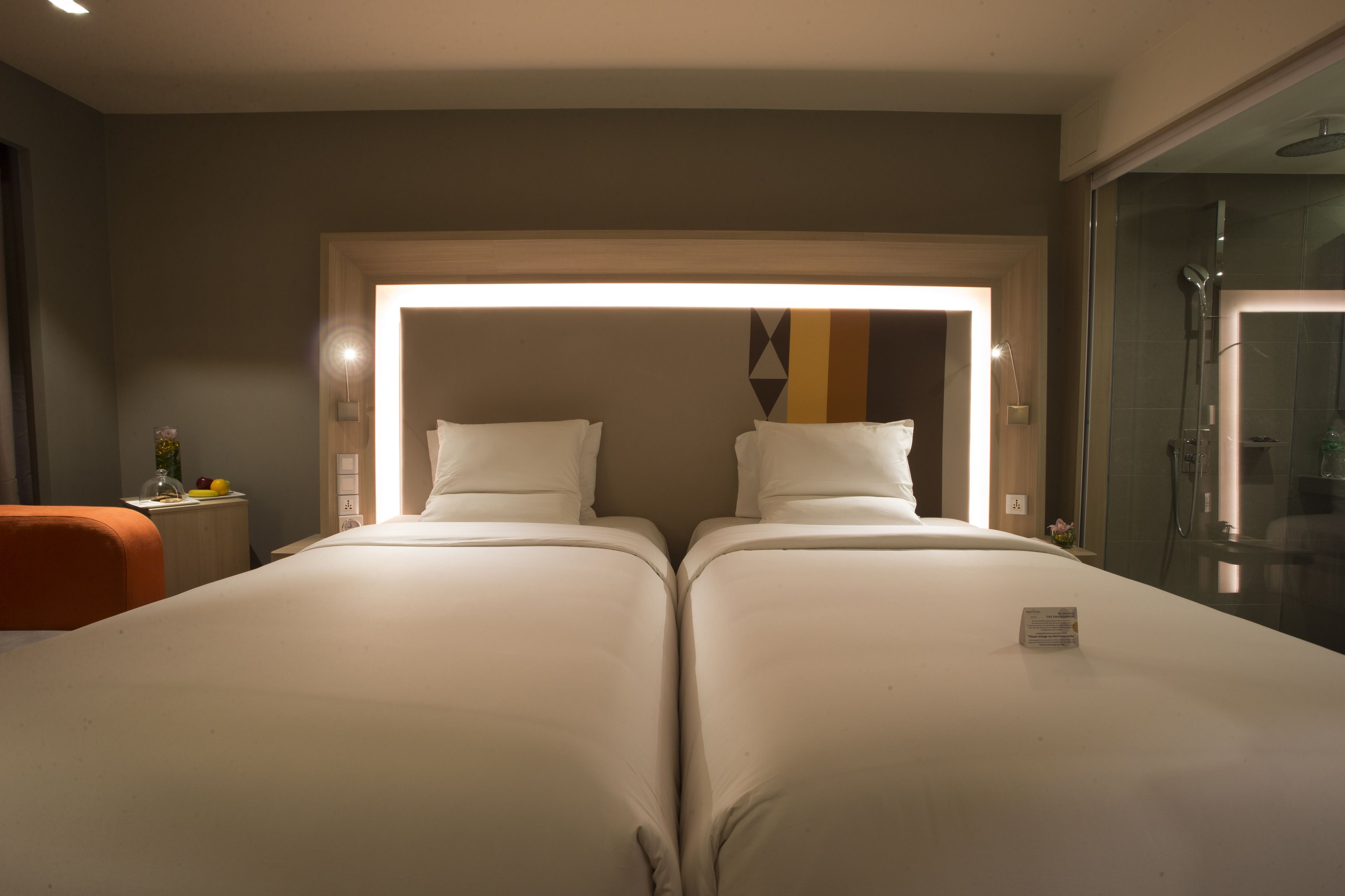 Une chambre d’hôtel avec deux lits, un espace douche et une petite table ornée d’un vase et de fruits.