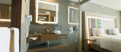 A modern hotel bathroom with a glass shower enclosure, a built-in sink, and a bedside table with a lamp.