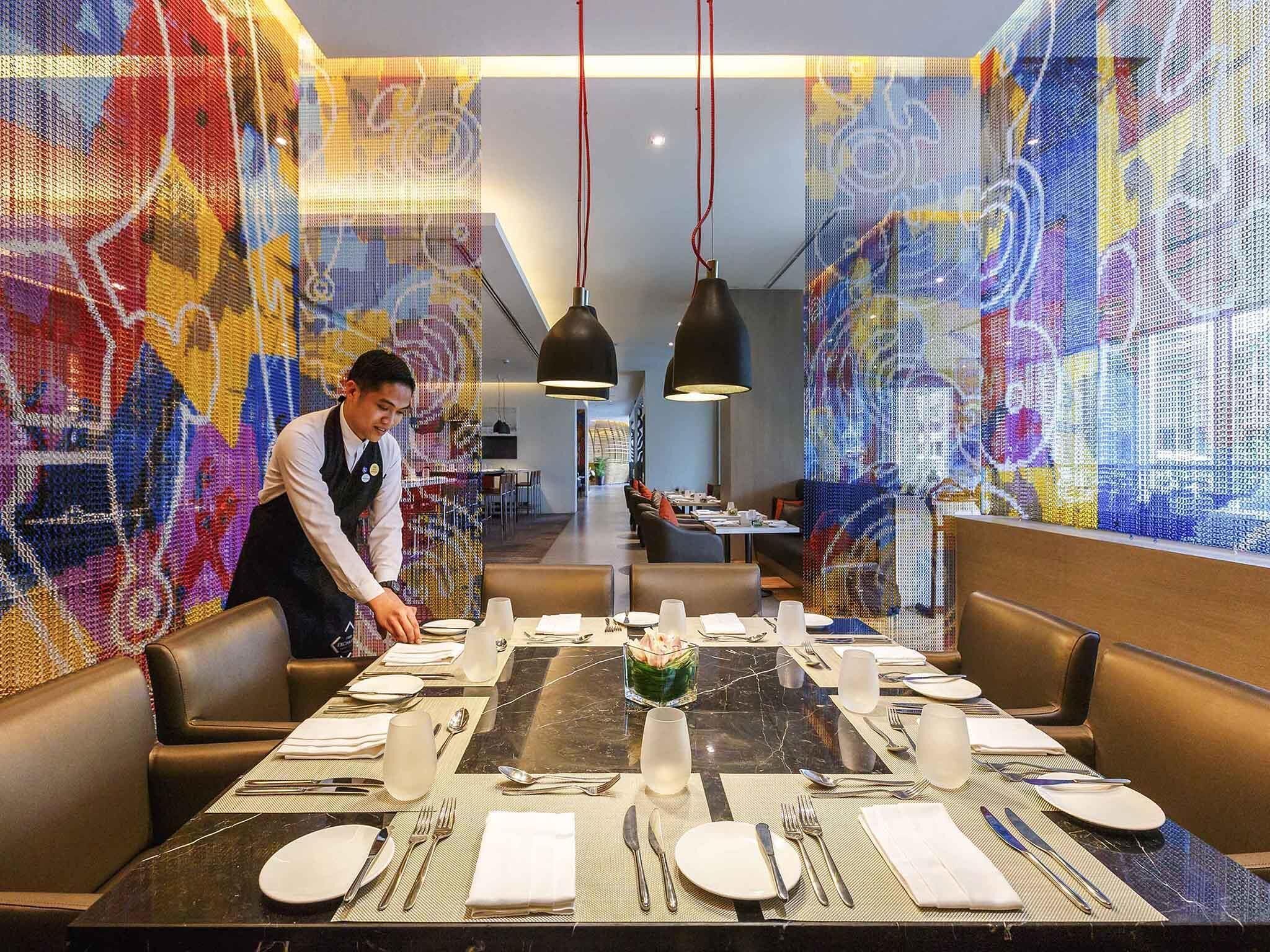 A restaurant with a long dining table set for a meal, a waiter attending to the table, and colorful abstract wall art.