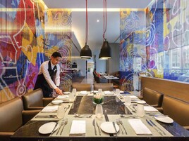Un restaurante con una larga mesa de comedor dispuesta para una comida, un camarero atendiendo la mesa, y coloridas obras de arte abstracto en las paredes.