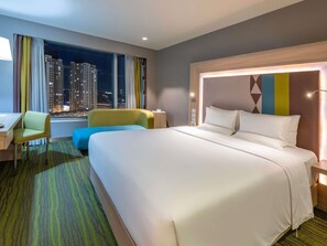 A hotel room with a large bed, a desk, a chair, and a view of a cityscape at night. - Novotel Manila Araneta City Hotel (Quezon City)