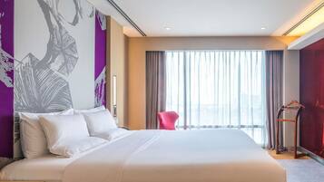 A hotel room with a large bed, a chair, a coat rack, and a view of the city through sheer curtains.