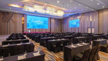 Une grande salle de conférence avec de nombreuses rangées de tables et de chaises, un grand écran affichant la mention « RÉUNION AU NOVOTEL », et un lustre moderne.