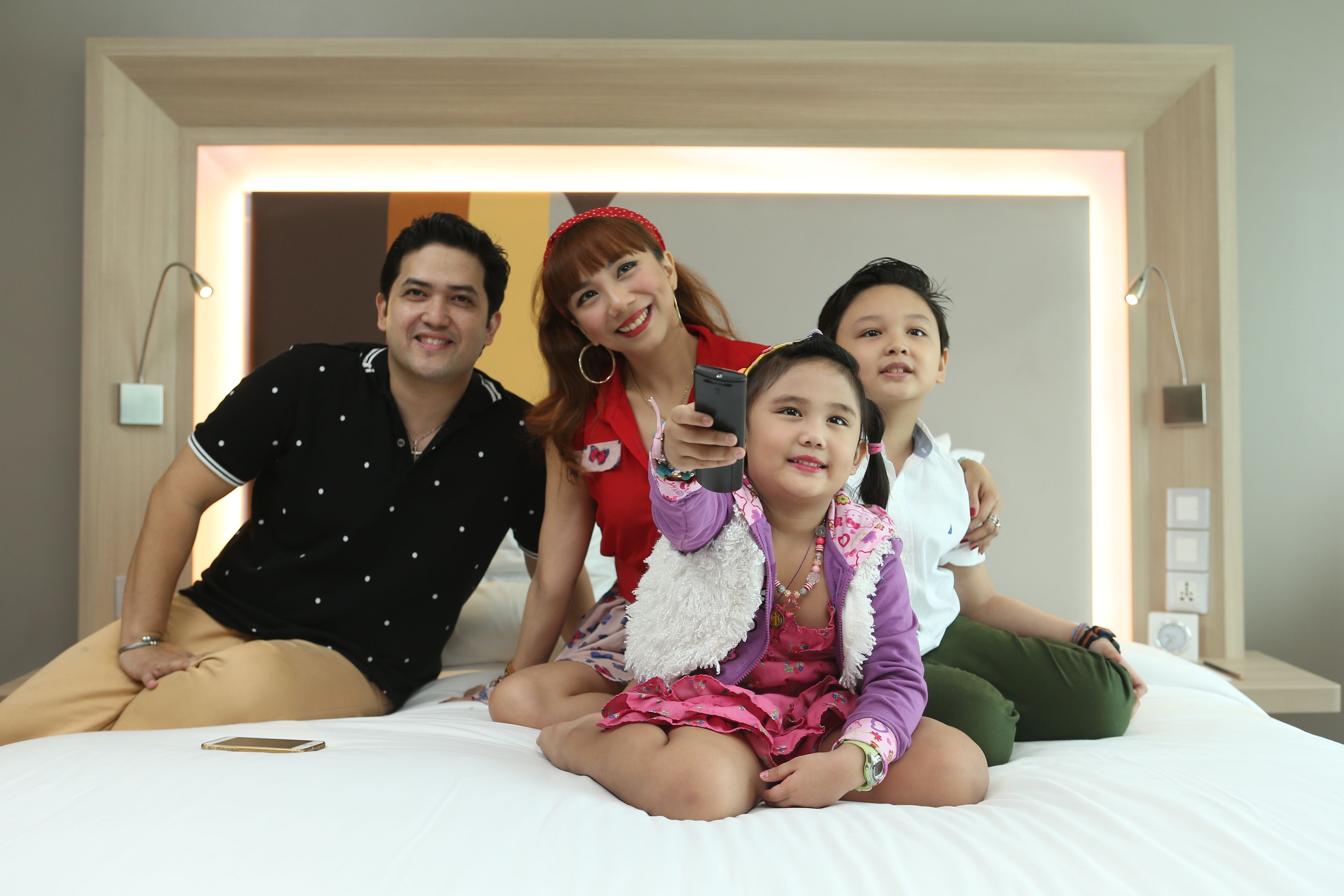 A group of people sitting on a bed in a hotel room with a modern design, including a nightstand and a lamp.