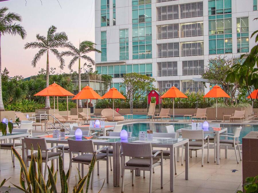 A poolside area with lounge chairs and tables, surrounded by palm trees and a modern building in the background.