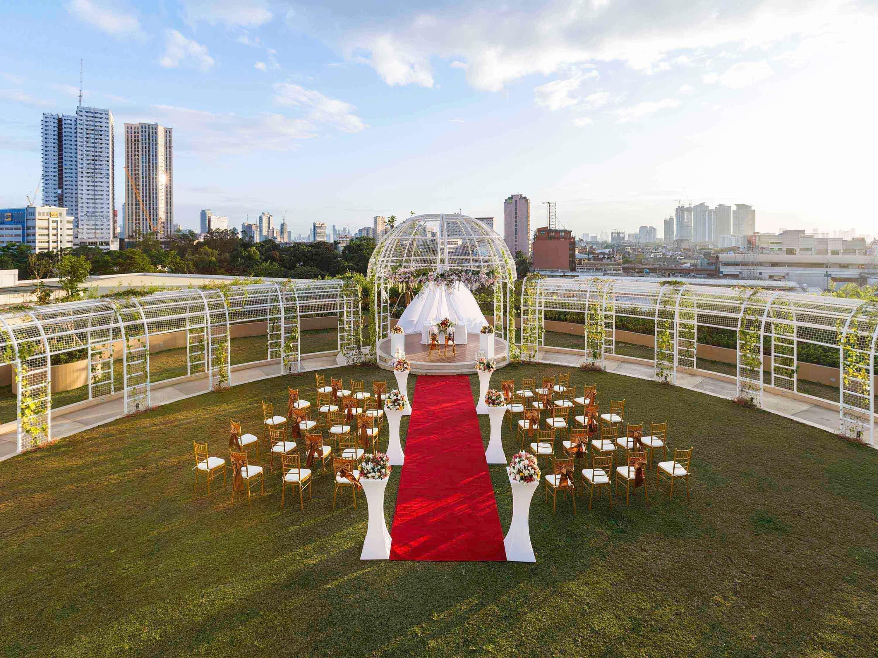 An outdoor wedding ceremony with a red carpet, white floral arrangements, and a city skyline in the background.