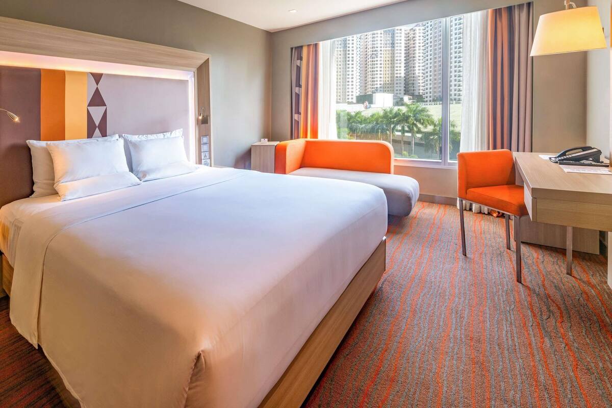 Une chambre d’hôtel avec un grand lit, un bureau, un fauteuil orange et une vue sur de hauts immeubles.